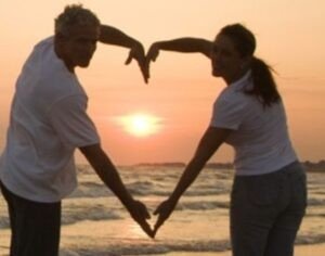 couple forming a heart with their hands in front of the beach at sunset