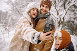 Parents take a winter selfie while their child stands aside, looking left out in the snow.
